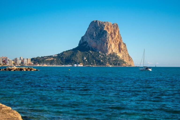 Impresionante paisaje marítimo del Peñon de Ifach con velero bajo un cielo azul claro en Calpe, España.